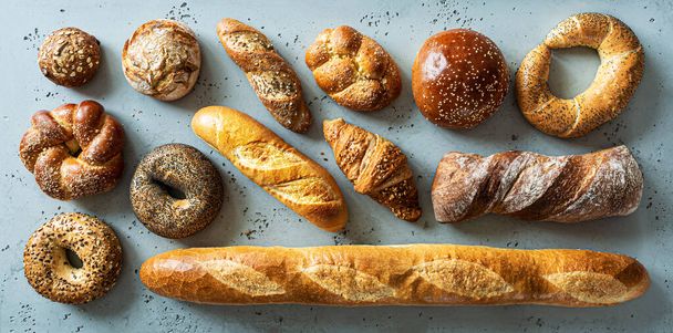 Bakery - various kinds of breadstuff. Bread rolls, baguette, bagel, burger bun and croissant captured from above (top view, flat lay). Grey stone background. - Foto, Imagen