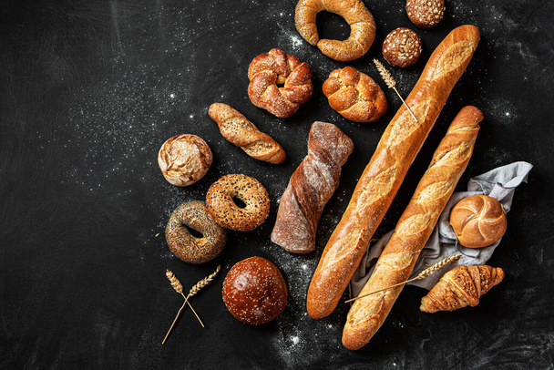 Bakery - various kinds of breadstuff. Bread rolls, baguette, bagel, burger bun and croissant captured from above (top view, flat lay). Black chalkboard background with free copy (text) space. - Fotó, kép