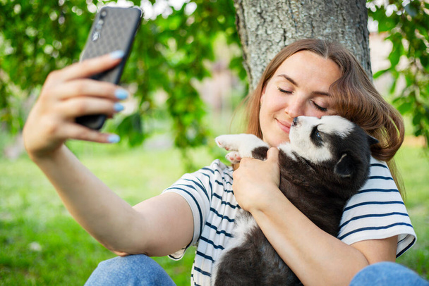 Young beautiful woman in a t-shirt and jeans with a husky puppy takes a photo or selfie on the phone while sitting in a park near a tree. - Zdjęcie, obraz