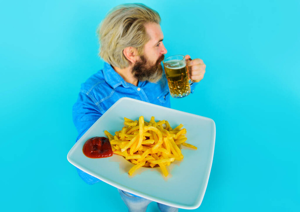 Bearded man with French fries on plate drinks beer. Snack at fast food. Selective focus. - Photo, Image