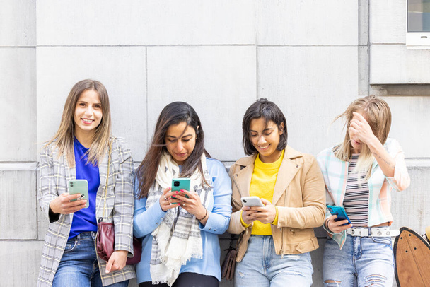 Grupo multiétnico de hermosas amigas jóvenes de pie contra una pared usando teléfonos inteligentes móviles - Foto, Imagen