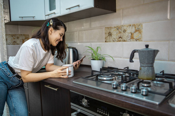 mujer beber té de café por la mañana en la cocina navegar por Internet en el teléfono comprobar los correos electrónicos - Foto, imagen