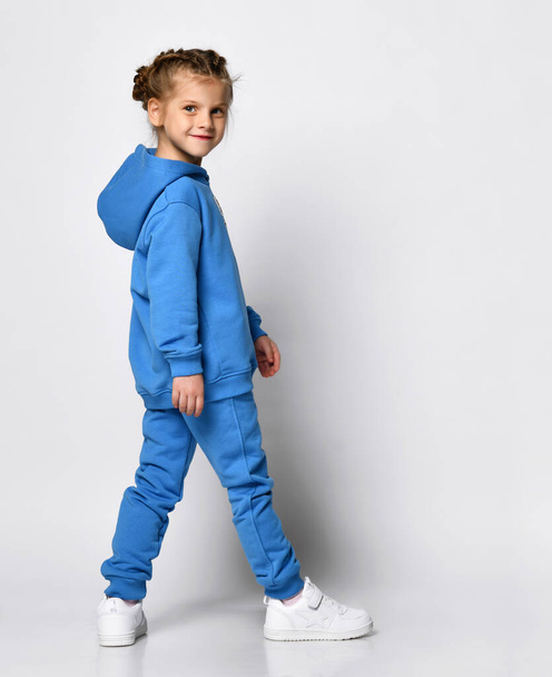 Active playful little girl in a trendy blue tracksuit having fun on a white background. A child with an emotional expression is standing half-sided and posing. Promotion of childrens sports fashion - Фото, изображение
