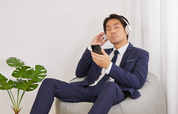 Asiatique homme d'affaires en costume tenir Smartphone porter casque pour écouter de la musique et s'asseoir sur le sac de haricot dans le bureau à domicile - Photo, image
