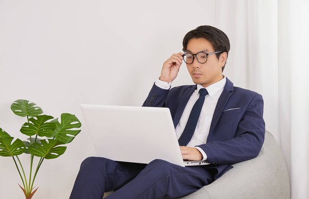 Ασιατικός οικονομικός σύμβουλος Αγγίξτε τα γυαλιά και χρησιμοποιήστε το lap-top στο εσωτερικό γραφείο. Επιχειρηματίας που εργάζεται στο σπίτι για την κοινωνική απόσταση ή κατ 'οίκον καραντίνα σε Covid 19 ή κατάσταση Coronavirus - Φωτογραφία, εικόνα