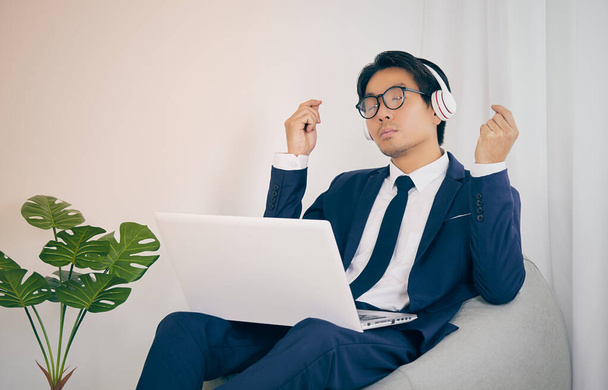 アジアのビジネスマンは、ヘッドフォンやメガネを着用ホームオフィスで音楽を聴く。ビンテージトーンのコヴィドやコロナウイルスの状況での社会的距離や家庭の隔離のためのリラックスした時間のビジネスマン - 写真・画像