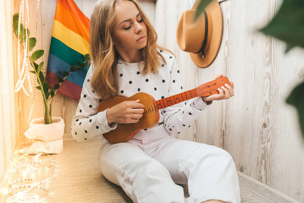 Νεαρή χίπισσα που κάθεται στο μπαλκόνι και παίζει κιθάρα. Μάθημα μουσικής και τραγουδιστής κάθεται. LGBTQ σημαία ουράνιο τόξο στο παρασκήνιο. Διαδικτυακά μαθήματα μουσικής. Παίζοντας όργανο και απολαμβάνοντας τραγούδια και αναψυχή - Φωτογραφία, εικόνα