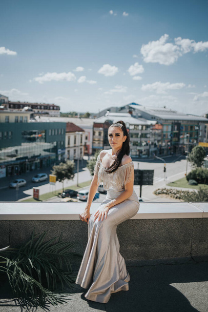 A vertical shot of a brunette female posing on a roof in a gorgeous beige evening dress - Фото, изображение