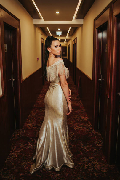 A vertical shot of a brunette female posing in a corridor in a gorgeous beige evening dress - Фото, изображение