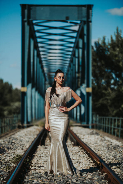 A shallow focus of a brunette woman wearing a luxurious sparkling dress and posing on old train tracks - Фото, изображение