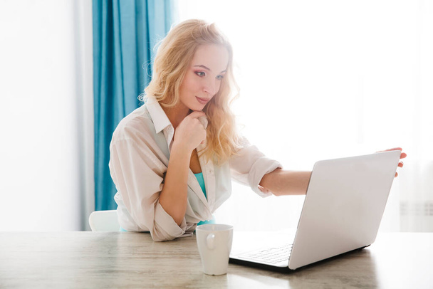 attractive young woman working with laptop in the kitchen, drinking coffee. home office. - Фото, изображение