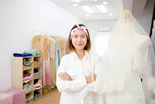 Los dueños de pequeñas empresas están trabajando felices en la sastrería. Una adolescente asiática trabaja como diseñadora de ropa, está midiendo y diseñando un vestido de novia sonriente. - Foto, imagen