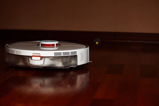 Έξυπνη σκούπα ρομπότ σε ξύλινο πάτωμα. Ρομπότ ηλεκτρική σκούπα εκτελεί αυτόματο καθαρισμό του διαμερίσματος σε ένα ορισμένο χρονικό διάστημα. Έξυπνο σπίτι - Φωτογραφία, εικόνα