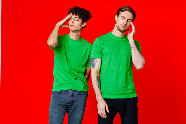 two friends in green t-shirts are standing next to tin with their hands emotions red background - Foto, Imagem