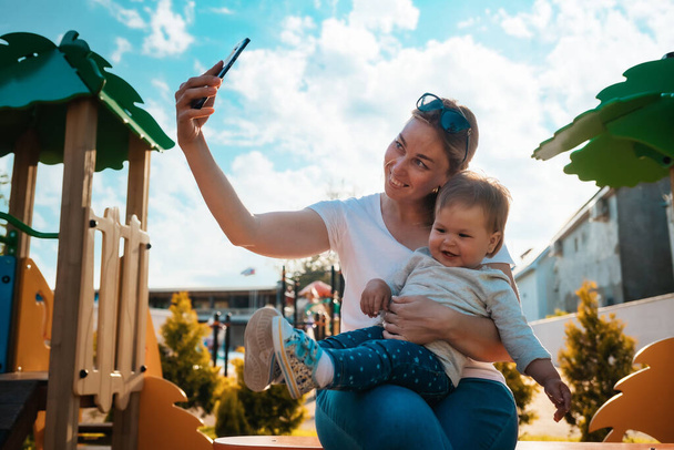Genç ve mutlu bir anne bebeği kollarında otururken selfie çeker. Arka planda oyun alanı. - Fotoğraf, Görsel