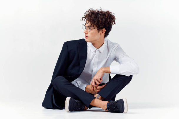 curly-haired guy in fashionable clothes and sneakers sits on the floor in a bright room in full growth - Фото, изображение