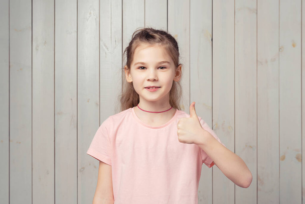 Fröhliches kleines Mädchen 9-11 Jahre zeigt Daumen hoch und sagt ja, lächelt zufrieden, genehmigt und mag etwas Gutes, lobt Wahl, steht in lässigem rosa T-Shirt über Holzgrund - Foto, Bild