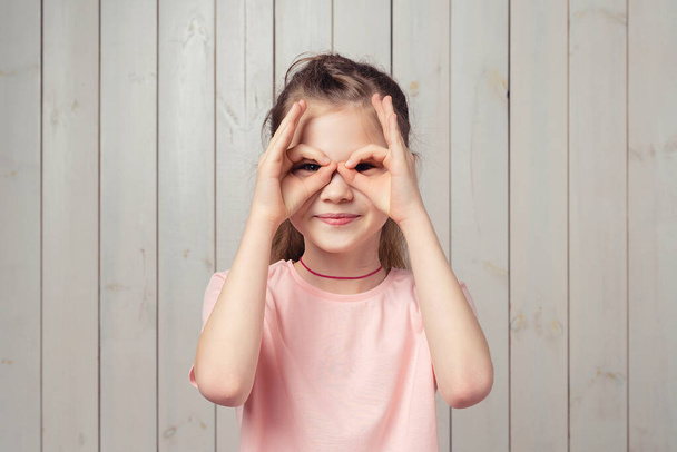 Bambina divertente 9-11 anni che tiene le dita vicino agli occhi come occhiali: Maschera come SuperHero, in piedi in casual t shirt rosa su sfondo di legno. Mostrando Ok Gesto - Foto, immagini