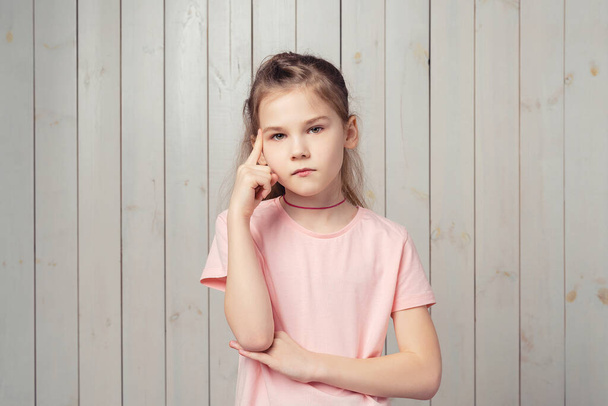 Thoughtful little girl 9-11 years old deep thinking, looking at camera with pondering face, making choice, standing pensive against wooden background - Foto, imagen