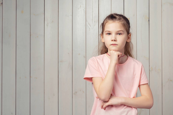 Głęboko myślę. Poważna dziewczynka 9-11 lat w luźnej różowej koszuli wyglądająca troskliwie, dotykająca podbródka i zastanawiająca się, podejmująca decyzję, stojąca nad drewnianym tłem - Zdjęcie, obraz