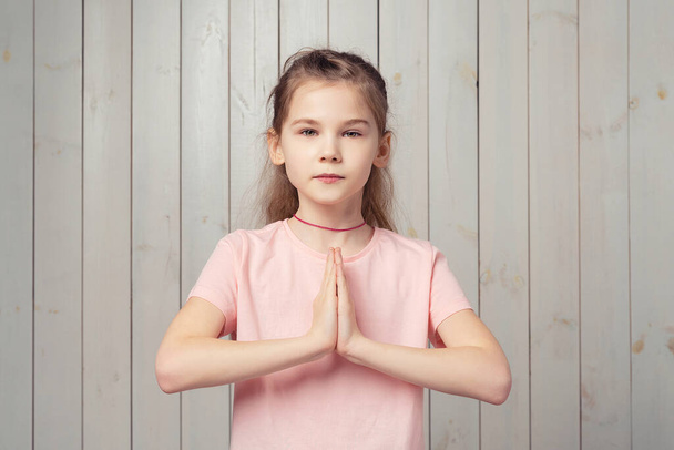 Upejpavá holčička 9-11 let v neformálním růžovém tričku prosí o laskavost, prosí, prosí rukama v modlícím se gestu, mračí se a vypadá smutně, stojí na dřevěném pozadí - Fotografie, Obrázek