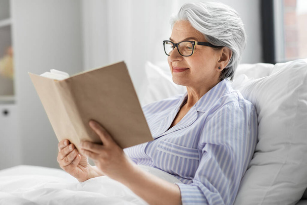 old woman in glasses reading book in bed at home - Foto, Bild