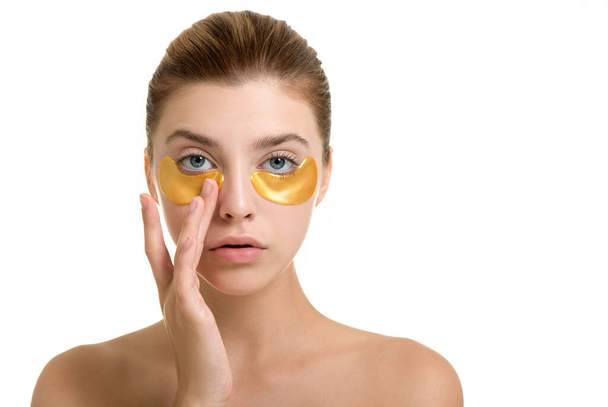 Beautiful young woman applies gold eye mask, isolated on white. Portrait of beauty model with natural nude make up cares about her skin. Spa, skincare and wellness. - Foto, Imagen