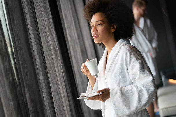 Beautiful woman relaxing at luxury hotel spa wearing bathrobe looking at window and drinking coffee - Fotó, kép
