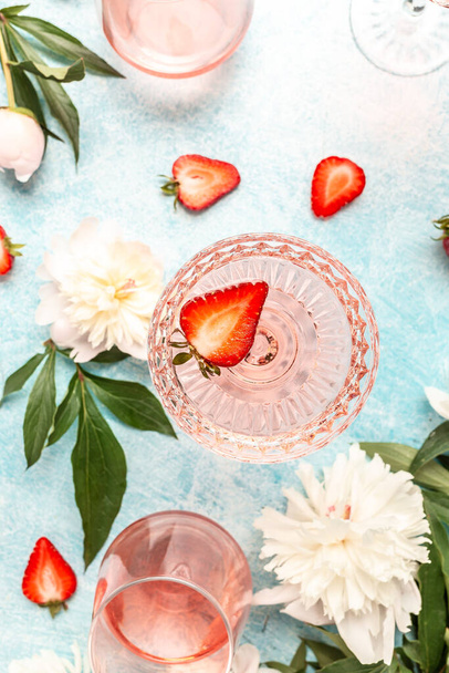 Lahodné růžové víno ve sklenicích s pivoňkovým květem a sladkými čerstvými jahodami na modrém pozadí, Letní nápoj pro párty, vinotéku nebo degustační koncept. - Fotografie, Obrázek