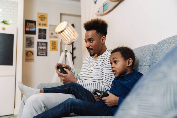 Focused african american father and son playing video game while sitting on couch at home - Foto, Imagen