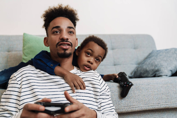 Focused african american father and son playing video game while sitting on couch at home - Φωτογραφία, εικόνα