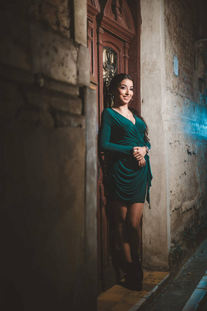 An adult woman wearing an elegant green dress and leaning on a building wall at night - Foto, afbeelding