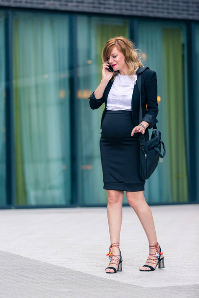 Uma mulher de negócios grávida caucasiana sorridente que está na rua em frente ao edifício de arquitetura moderna enquanto fala em seu telefone celular. Ela está segurando um saco de laptop. - Foto, Imagem