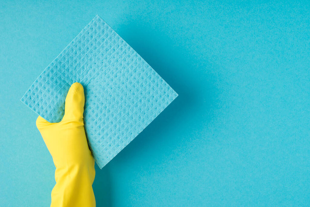 Top view photo of hand in yellow rubber glove holding blue viscose rag on isolated pastel blue background with copyspace - Photo, Image