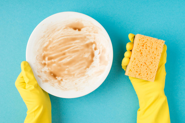 Top view photo of hands in yellow rubber gloves holding dirty plate and scouring pad on isolated pastel blue background - Photo, Image