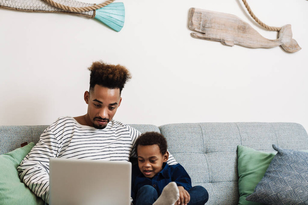 Focused african american father and son hugging and using laptop while sitting at home - Foto, Imagem