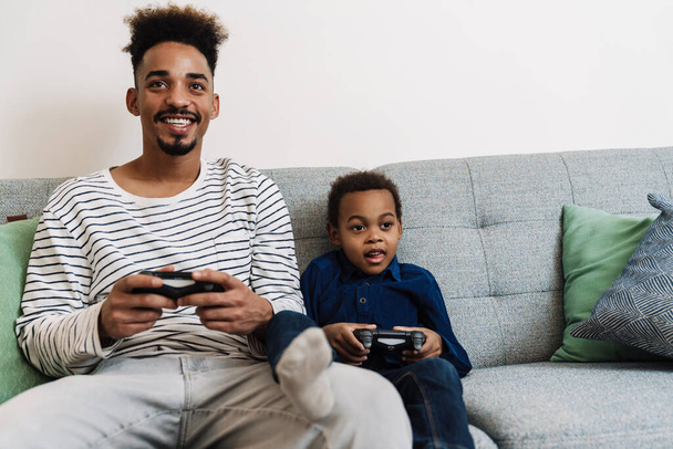 Happy african american father and son playing video game while sitting on couch at home - Photo, Image
