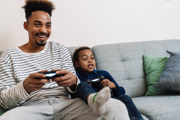 Happy african american father and son playing video game while sitting on couch at home - 写真・画像