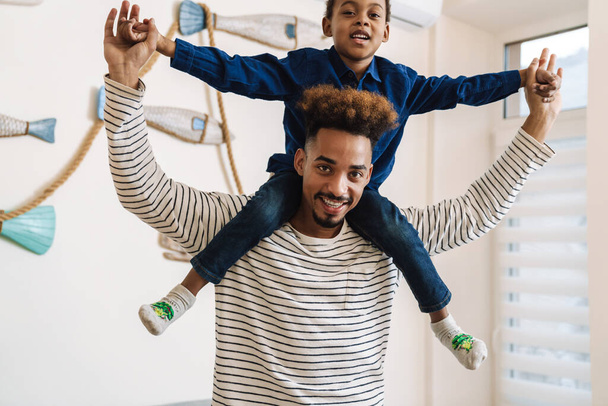 Happy african american boy having fun and sitting on neck of his smiling father at home - Foto, imagen