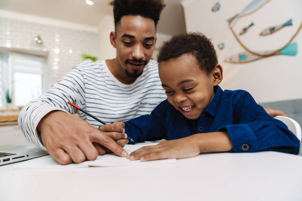 Blij Afrikaans-Amerikaanse vader doet huiswerk met zijn gelukkige zoon thuis - Foto, afbeelding