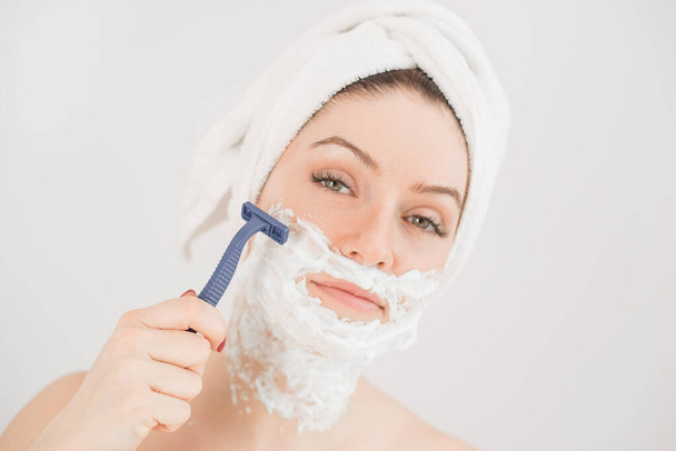 Cheerful caucasian woman with a towel on her head and shaving foam on her face holds a razor on a white background - Foto, imagen
