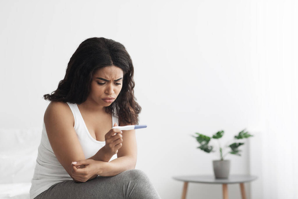 Donna afroamericana sconvolta che controlla il suo recente test di gravidanza, seduta sul letto a casa, spazio libero - Foto, immagini