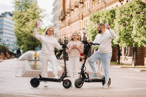 A family in white clothes stands in the city on electric scooters and drinks a drink. - Фото, изображение