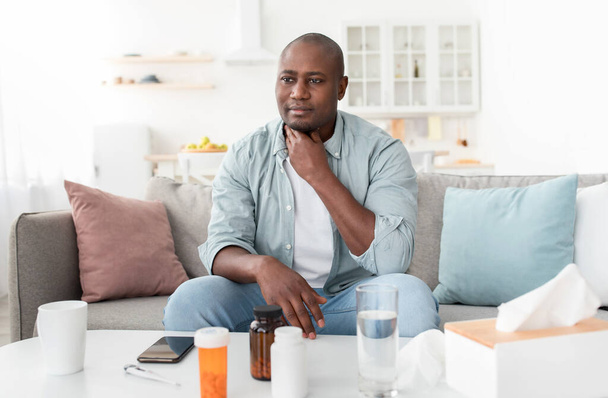 Starší africký Američan se cítí špatně, trpí bolestí v krku, sedí na pohovce u stolu s ubrousky a prášky - Fotografie, Obrázek