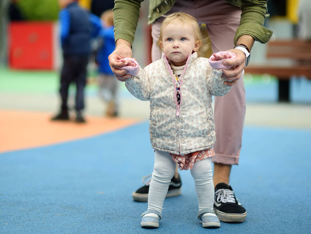 Cute toddler girl having fun on outdoor playground. Young father is teaching his little daughter to walk. First step of baby. Walking for family with little kids. Happy parenthood. - Fotografie, Obrázek