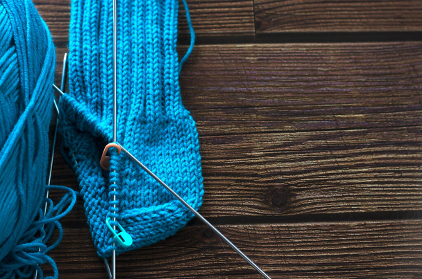 Πλέκοντας μια ζεστή κάλτσα από μπλε μάλλινο νήμα σε βελόνες πλεξίματος. Πλέξιμο του έργου σε εξέλιξη. Καφέ ξύλινο τραπέζι. Άνω όψη. - Φωτογραφία, εικόνα