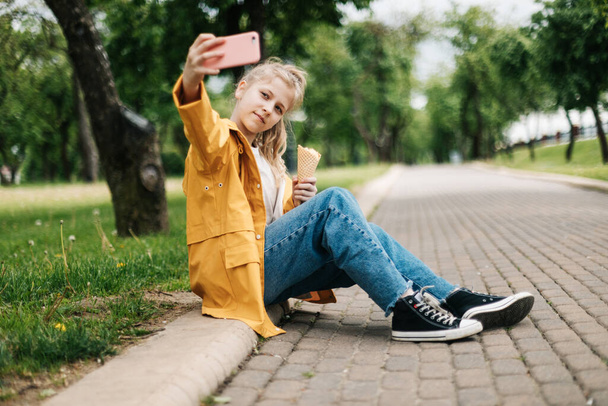 Yolda oturan bir sarışın, elinde telefon ve dondurmayla komik bir selfie çekiyor. Çocuk dışarıda. - Fotoğraf, Görsel