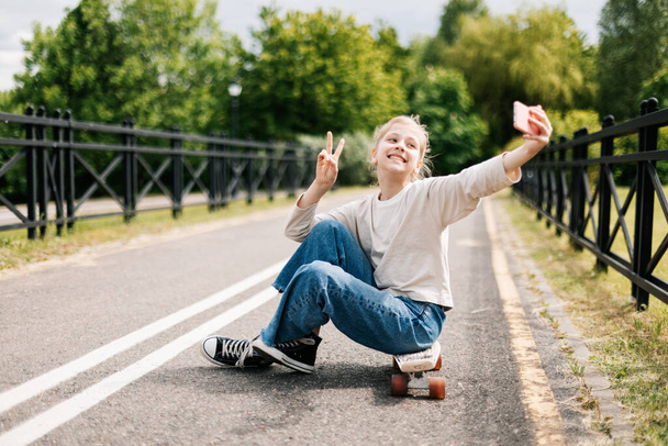 Милая блондинка-подросток сидит на скейтборде в городском парке, болтает на смартфоне с друзьями и делает селфи.. - Фото, изображение