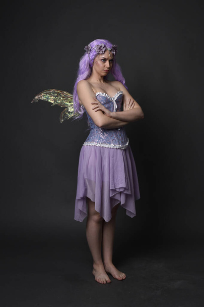 Teljes hosszúságú portré egy lila hajú lányról, aki fantázia fűzőt visel tündér szárnyakkal és virágkoronával. Álló póz egy sötét stúdió háttér. - Fotó, kép