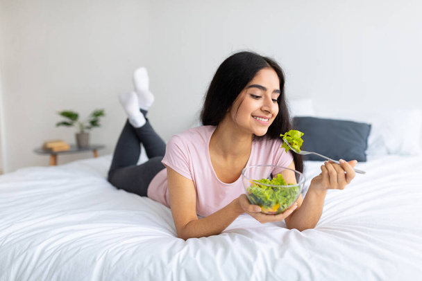 Сбалансированная сырая пища. Прекрасная индийская женщина ест свежий овощной салат, лежит на кровати дома с вкусной едой - Фото, изображение
